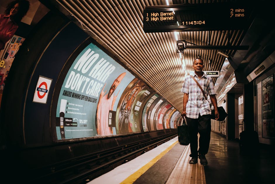 A rectangular black and white street sign mounted on a black pole, indicating directions to Arsenal Station with an upward arrow, situated above dense foliage including trees with green and reddish leaves. The sign features the London Underground roundel logo with the blue bar displaying 'Arsenal Station.' In the background, the sky is partly cloudy with patches of blue visible. The surrounding environment suggests an urban area in Islington, with rooftops and chimney stacks partly visible below the sign, supporting the context of house removals and access issues on narrow streets. The sign is positioned at a height, supported by additional brackets, and is surrounded by the natural elements and typical London street features that are relevant to relocation logistics and transportation planning by Islington Movers.