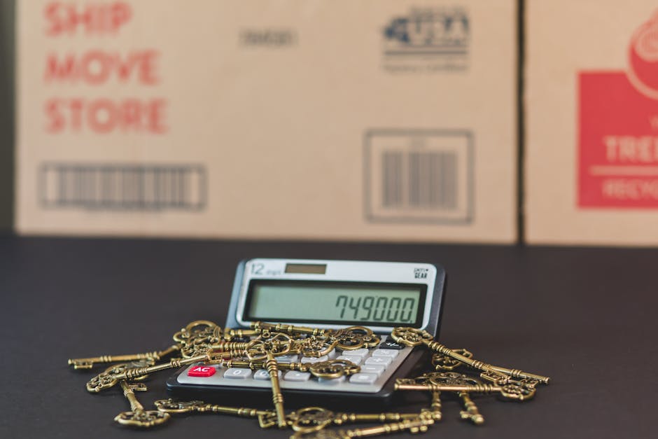 A close-up view of a calculator displaying the number 749800, surrounded by a scattered pile of antique-style gold keys on a black surface. In the background, there are partially visible brown cardboard boxes with red and black printed labels, one listing options like 'ship,' 'move,' and 'store,' indicating a home relocation or moving scenario. The scene is indoors with soft lighting, capturing elements typical of furniture transport, packing, and loading processes associated with house removals, as performed by professional services such as Islington Movers, which specialize in removals in the Islington area.