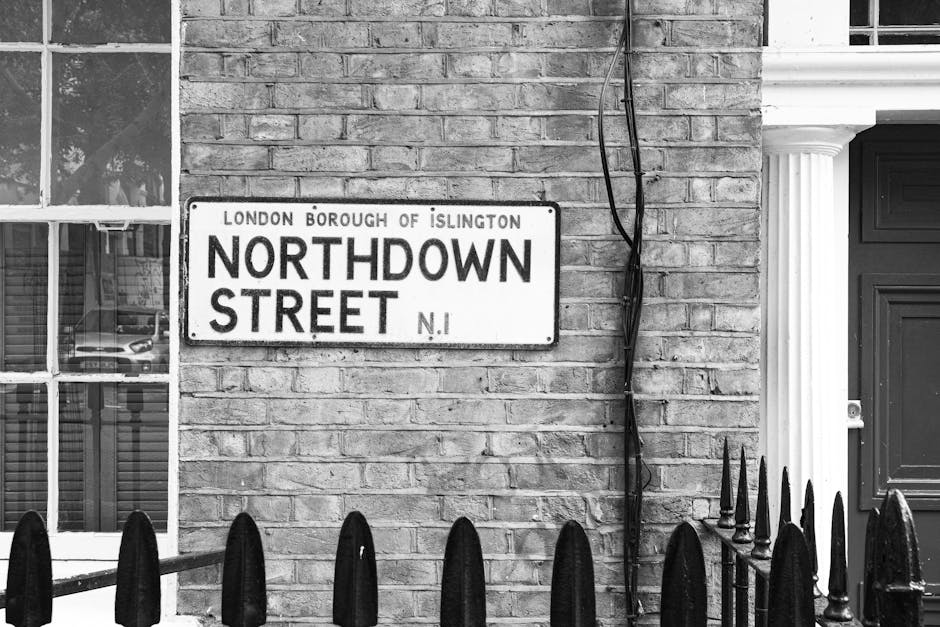 A black and white photograph of a residential building's exterior showing a street sign reading 'Northdown Street, London Borough of Islington' mounted on a brick wall. The wall features a window with multiple panes on the left side, partially obscured by a black metal railing in the foreground. To the right of the sign, a black cable runs vertically along the brick surface, secured with clips. The building has a classic architectural style with a decorative white column and a black door with a mail slot below a small ornamental window. The scene suggests an urban setting suitable for house removals, with visible elements such as the building facade and street sign providing context for location. Islington Movers, a professional removals company, may use such imagery to illustrate aspects of home relocation and furniture transport in the Islington area, supporting their services involved in loading and moving household items in urban environments.