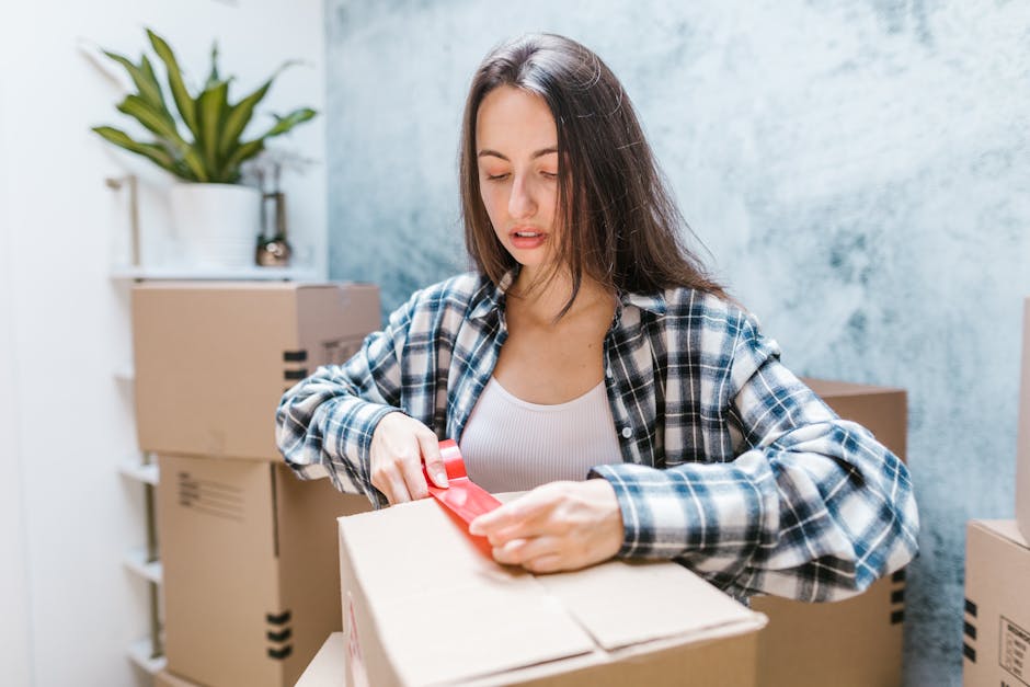 A woman with long brown hair, wearing a blue and white plaid shirt over a white top, is seen in an indoor setting surrounded by several cardboard moving boxes, some sealed with packing tape. She appears to be fastening or adjusting a red strap or ribbon on one of the boxes, which are stacked in the background on a white shelving unit and on the floor. A potted plant on the top shelf provides greenery against a light blue, textured wall, while natural light illuminates the scene. This image depicts the packing stage of a home relocation process, highlighting the careful handling and packing of belongings during furniture transport or removal services. It reflects the services offered by Islington Movers, focusing on efficient and organised packing for residential moves, and illustrates the logistics involved in loading and preparing items for transport during a house removal or moving company’s operations.
