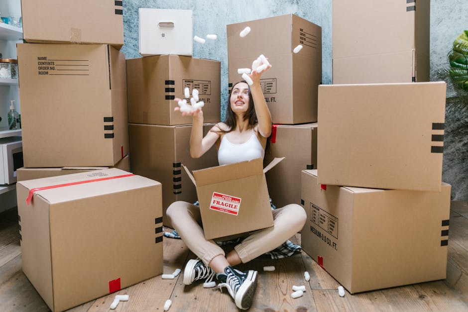 A young woman with long dark hair and casual attire, including a white tank top, beige pants, and sneakers, sits cross-legged on a wooden floor inside a room filled with large cardboard moving boxes of various sizes, some sealed with red tape and others with black markings. She is smiling and tossing packing material, such as white foam packing peanuts, into the air above her head. One of the boxes in front of her has a red and white 'Fragile' label attached, indicating careful handling. The boxes are stacked and arranged around her, with some placed against a wall that features a textured light blue and grey finish. Nearby, a small green plant and a glimpse of a kitchen countertop are visible, suggesting a residential setting during a home relocation process. This scene illustrates packing and loading activities commonly associated with furniture transport and home moves, with priority on organizing belongings before the moving day. Islington Movers provides professional help with safe packing and efficient removals to ensure smooth home relocations in the Islington area.