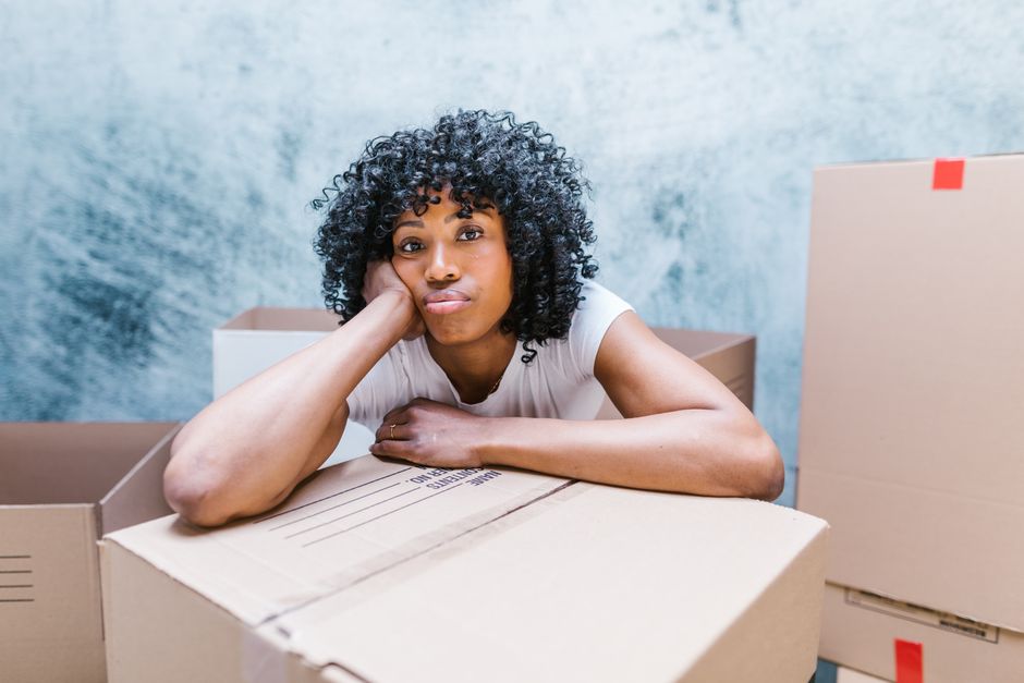 A woman with long brown hair, wearing a blue and white plaid shirt over a white top, is seen in an indoor setting surrounded by several cardboard moving boxes, some sealed with packing tape. She appears to be fastening or adjusting a red strap or ribbon on one of the boxes, which are stacked in the background on a white shelving unit and on the floor. A potted plant on the top shelf provides greenery against a light blue, textured wall, while natural light illuminates the scene. This image depicts the packing stage of a home relocation process, highlighting the careful handling and packing of belongings during furniture transport or removal services. It reflects the services offered by Islington Movers, focusing on efficient and organised packing for residential moves, and illustrates the logistics involved in loading and preparing items for transport during a house removal or moving company’s operations.