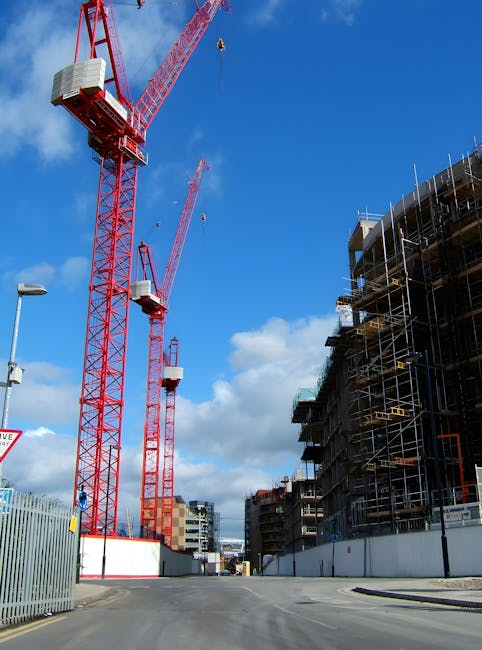 Photograph of a construction site on a city street under a bright blue sky with scattered clouds, showing two large red tower cranes with white counterweights positioned next to a partially completed multi-story building surrounded by metal scaffolding and safety barriers. The cranes are lifting equipment or materials high above the ground, with the longer jib of the crane extending horizontally. In the foreground, there is an empty paved road with a lamppost on the left side and a metal fence running along the street, indicating an active construction and development zone. This scene reflects urban building and development processes, which are part of the broader infrastructure and housing growth relevant to house removals and relocation services, such as those offered by Islington Movers.