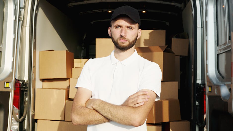 A young male mover with a beard, wearing a white polo shirt and black cap, standing inside the open rear of a moving van with its doors wide open. Behind him, the van is filled with neatly stacked cardboard boxes of various sizes, some wrapped in plastic or paper for protection. The boxes are arranged in an organized manner on the vehicle's floor, indicating an ongoing home relocation process. The van is situated on a paved surface, with sunlight illuminating the interior and the surrounding area. The image reflects a professional furniture transport and packing process typical of house removal services, with [COMPANY_NAME] handling the loading of household items for a timely and efficient move.