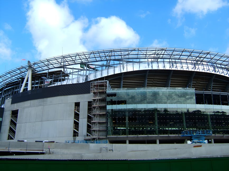 Photograph of a construction site on a city street under a bright blue sky with scattered clouds, showing two large red tower cranes with white counterweights positioned next to a partially completed multi-story building surrounded by metal scaffolding and safety barriers. The cranes are lifting equipment or materials high above the ground, with the longer jib of the crane extending horizontally. In the foreground, there is an empty paved road with a lamppost on the left side and a metal fence running along the street, indicating an active construction and development zone. This scene reflects urban building and development processes, which are part of the broader infrastructure and housing growth relevant to house removals and relocation services, such as those offered by Islington Movers.