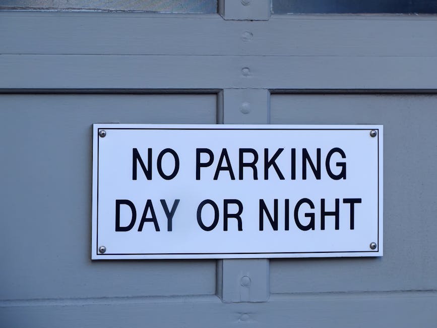 A rectangular white sign with bold black lettering stating 'NO PARKING DAY OR NIGHT' is mounted on a grey wooden door or panel, secured with four screws, two in the top corners and two in the bottom corners. The background consists of horizontal and vertical grey wooden planks, possibly part of a garage or building exterior. The lighting is neutral, indicating an outdoor setting. This sign is relevant to house removals and furniture transport, as it indicates parking restrictions that may affect loading and unloading processes during home relocation, which is a key consideration in planning moves according to Islington Council parking permit rules for removals, as explained by Islington Movers.