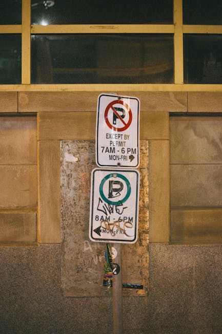 Close-up of a vertical parking restriction and permit zone sign mounted on a metal pole against a stone wall, beneath a large window with yellow framing. The upper part of the sign displays a 'No Parking' symbol with text indicating parking restrictions from 7 am to 6 pm, Monday to Friday, with an exemption for permit holders. The lower part shows a 'Permit Holders Only' symbol with additional details about parking restrictions from 8 am to 6 pm, Monday to Friday. Behind the signs, the wall consists of beige stone panels, and the window above features a dark-tinted glass with yellow framing. The scene suggests a location where moving and removal services, such as those provided by Islington Movers, need to comply with local parking regulations during home relocation or furniture transport in Islington.