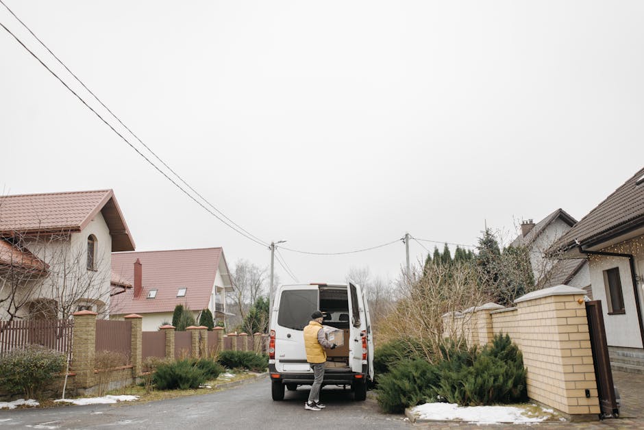 A man wearing a yellow and black jacket is loading a cardboard box into the rear of a white cargo van parked on a residential street. The van's rear doors are open, revealing a spacious interior suitable for home relocation and furniture transport. The van is positioned on the paved road next to a sidewalk, with some low shrubs and patches of snow visible on the ground. In the background, standalone houses with red-tiled roofs, brick and stucco walls, and garden fencing line the street. Overhead, power lines run parallel to the street, and the sky is overcast, indicating a cloudy day. Islington Movers' professional removals service is implied by the context of the loading process, demonstrating a typical scene of packing, loading, and preparing for a local move within the Barnsbury estates area.
