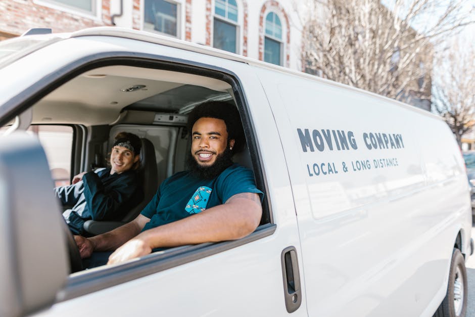 Inside a white moving van with the sliding door open, two movers from Islington Movers are visible. The man in the foreground, seated in the driver’s seat, has a dark beard, short curly hair, and is smiling at the camera, wearing a blue uniform with a logo on the sleeve. The woman in the background, seated in the passenger seat, has dark hair pulled back with a headband, and is also smiling. The interior of the van shows a cargo area with packing materials such as cardboard boxes and plastic-wrapped furniture, indicating preparation for a house removal or home relocation service. The van is parked outdoors on a city street with a residential building visible in the background, under natural daylight. The scene captures the loading process, with a focus on the team members ready to assist with furniture transport and packing and moving tasks, reflecting professional small-scale moving services offered by Islington Movers.