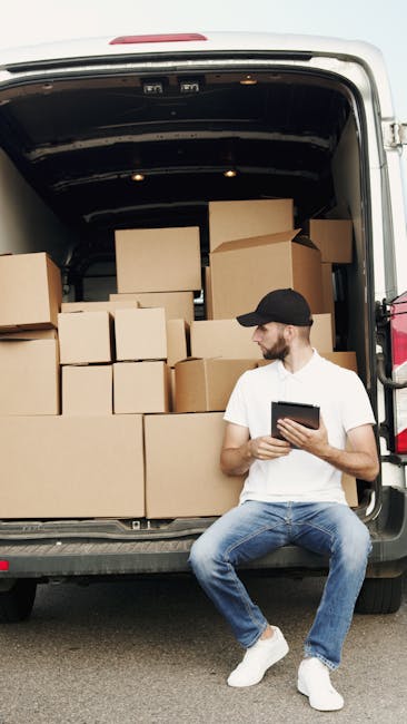 A man wearing a yellow and black jacket is loading a cardboard box into the rear of a white cargo van parked on a residential street. The van's rear doors are open, revealing a spacious interior suitable for home relocation and furniture transport. The van is positioned on the paved road next to a sidewalk, with some low shrubs and patches of snow visible on the ground. In the background, standalone houses with red-tiled roofs, brick and stucco walls, and garden fencing line the street. Overhead, power lines run parallel to the street, and the sky is overcast, indicating a cloudy day. Islington Movers' professional removals service is implied by the context of the loading process, demonstrating a typical scene of packing, loading, and preparing for a local move within the Barnsbury estates area.