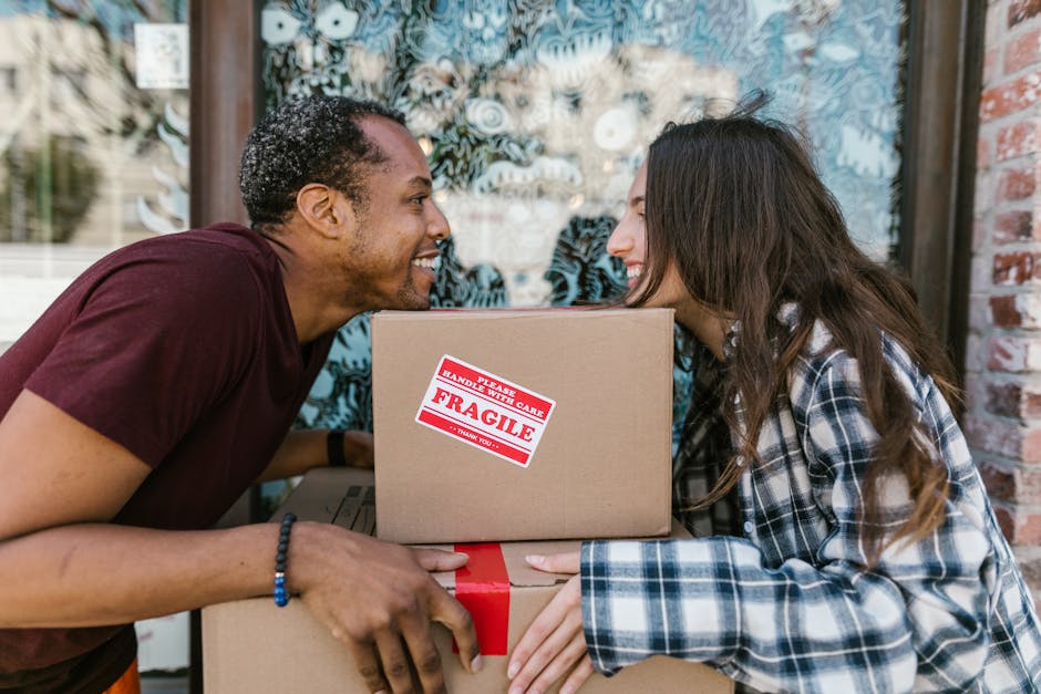 Two individuals engaged in a home relocation process outdoors, with a brick wall and a window with patterned curtains in the background. The man on the left, wearing a maroon T-shirt and a beaded bracelet, is smiling and leaning forward, holding a medium-sized cardboard box with a red and white 'Fragile' handling label. The woman on the right, dressed in a blue and white plaid shirt, is also smiling and facing the man, holding a larger cardboard box secured with red packing tape. The boxes, made of cardboard and covered with plastic wrap, are part of the loading process as they prepare to transport belongings for a move, which is characteristic of furniture transport and packing and moving services. The scene reflects a friendly interaction during a house removal or relocation, with the boxes positioned in front of them, suggesting they are in the midst of a moving day. Islington Movers, providing professional removals, often supports such home relocations, ensuring safe and efficient packing and transportation.