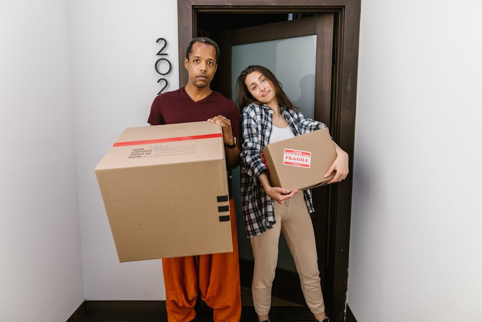 Two individuals, a man and a woman, are standing inside a residential doorway, each holding a cardboard box as part of a home relocation or furniture transport process. The man, dressed in an orange p